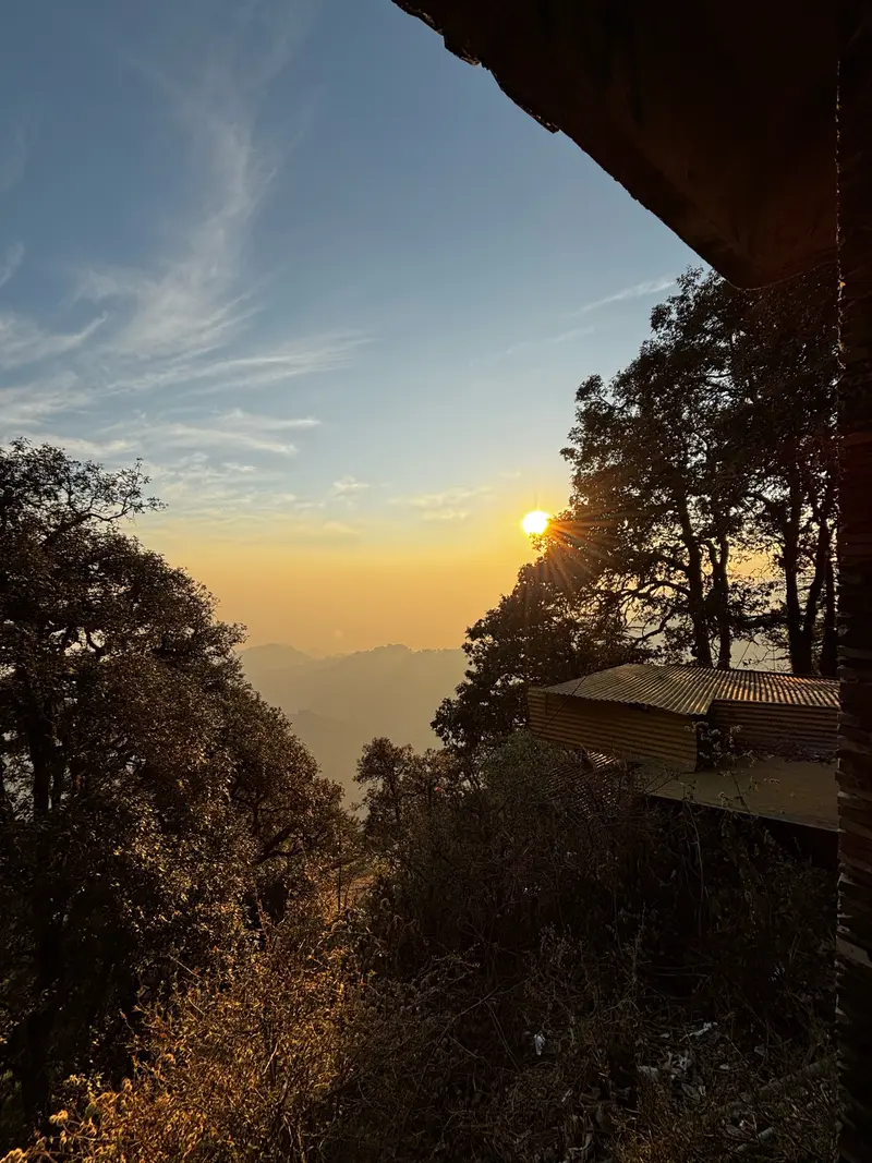 Golden sunset sunburst setting behind mountain trees and a hillside cabin in Mussoorie.