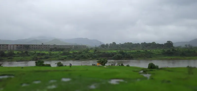 Panoramic landscape of a river and lush green hills under overcast monsoon skies.