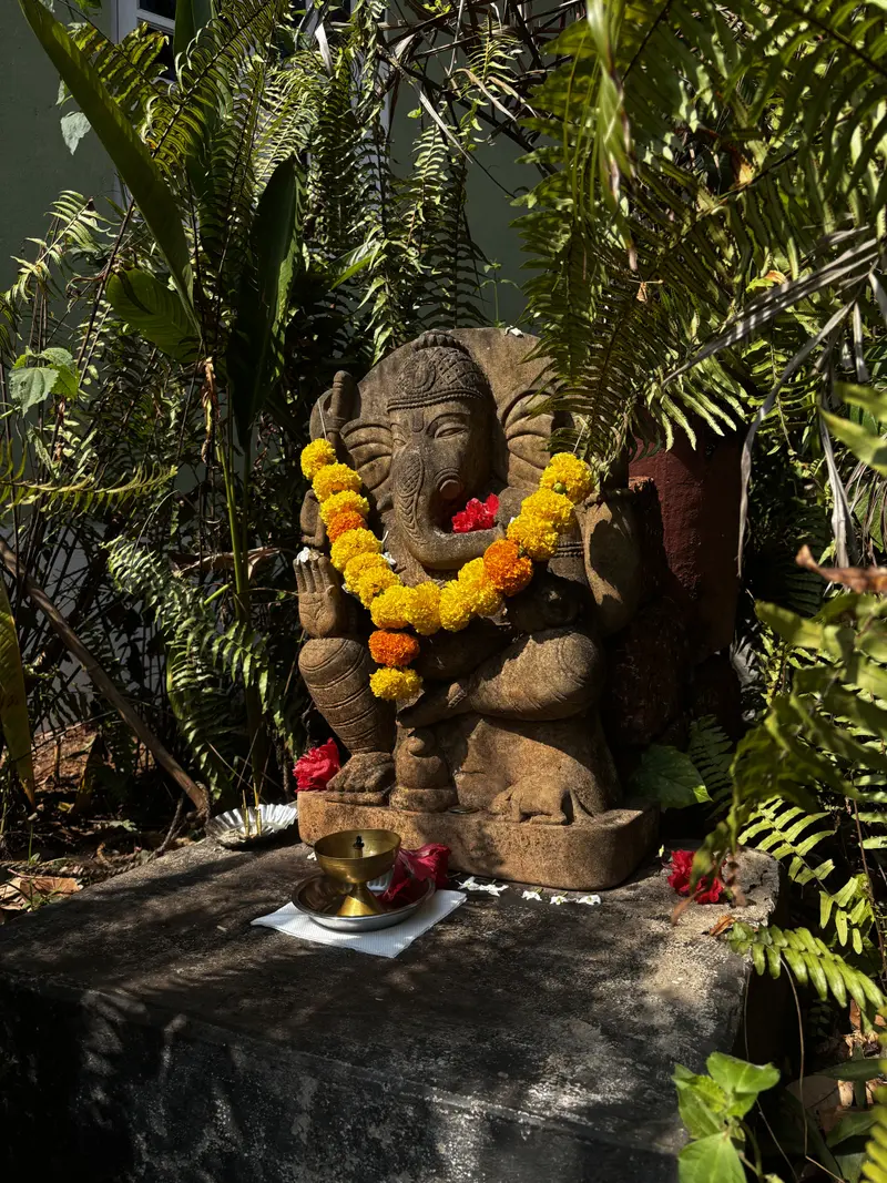 Stone idol of Lord Ganesha with yellow marigolds and a brass diya among green ferns.