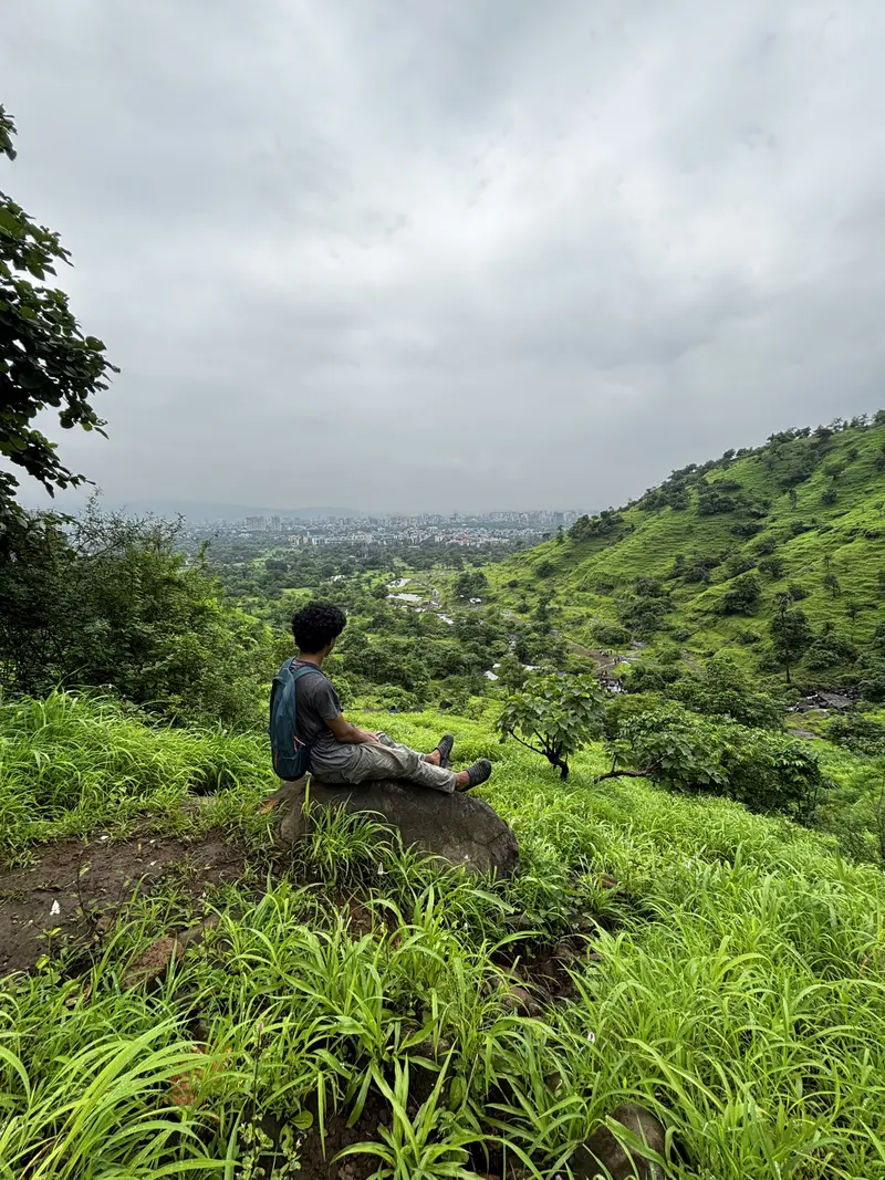 Heramb on a recent mountain trek, sitting on a rocky ledge overlooking green hills and a distant valley.